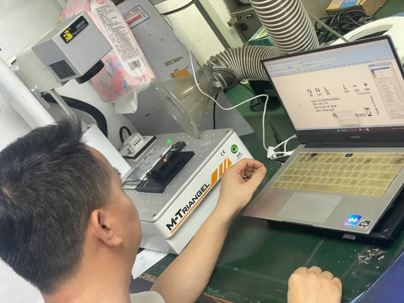 An engineering specialist calibrating a laser engraving machine and verifying parametric text on a laptop for smart switch enclosure labeling at the Shenzhen factory.